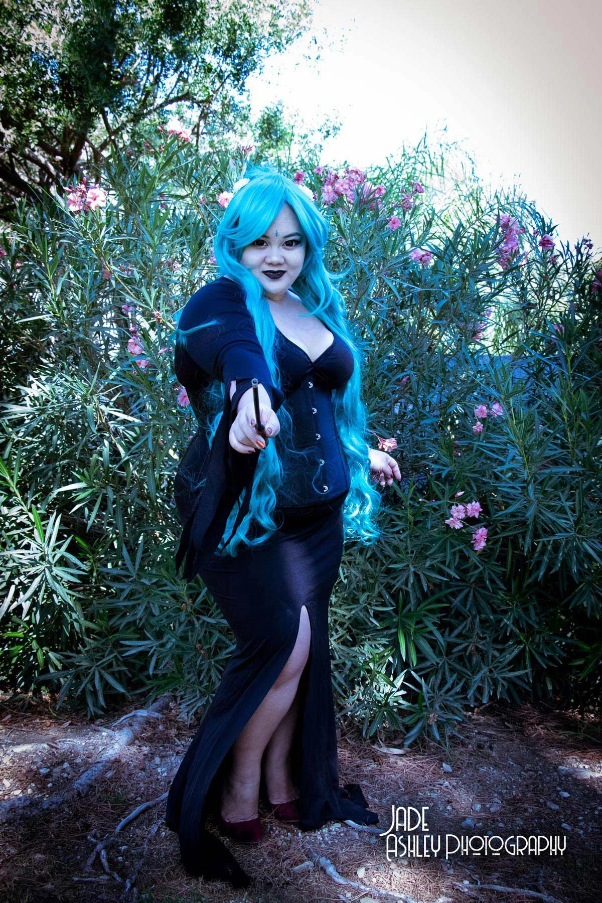 A woman dressed as a dark sorceress or witch with long blue hair, dark makeup, and a black dress with side slit, standing amid pink flowering bushes outdoors during daylight, pointing a wand towards the camera.
