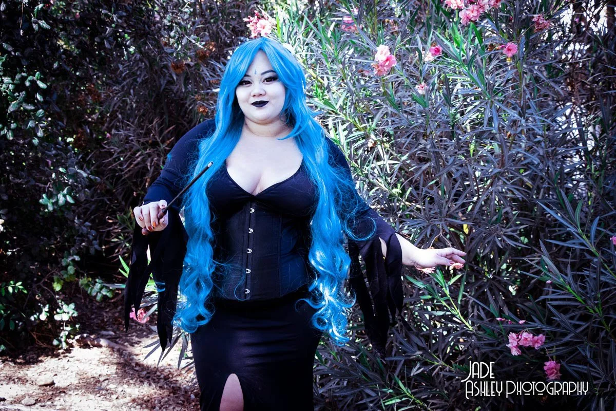 Woman with long blue hair dressed in black standing among purple and green foliage, holding a wand, with dark makeup and gothic style clothing.