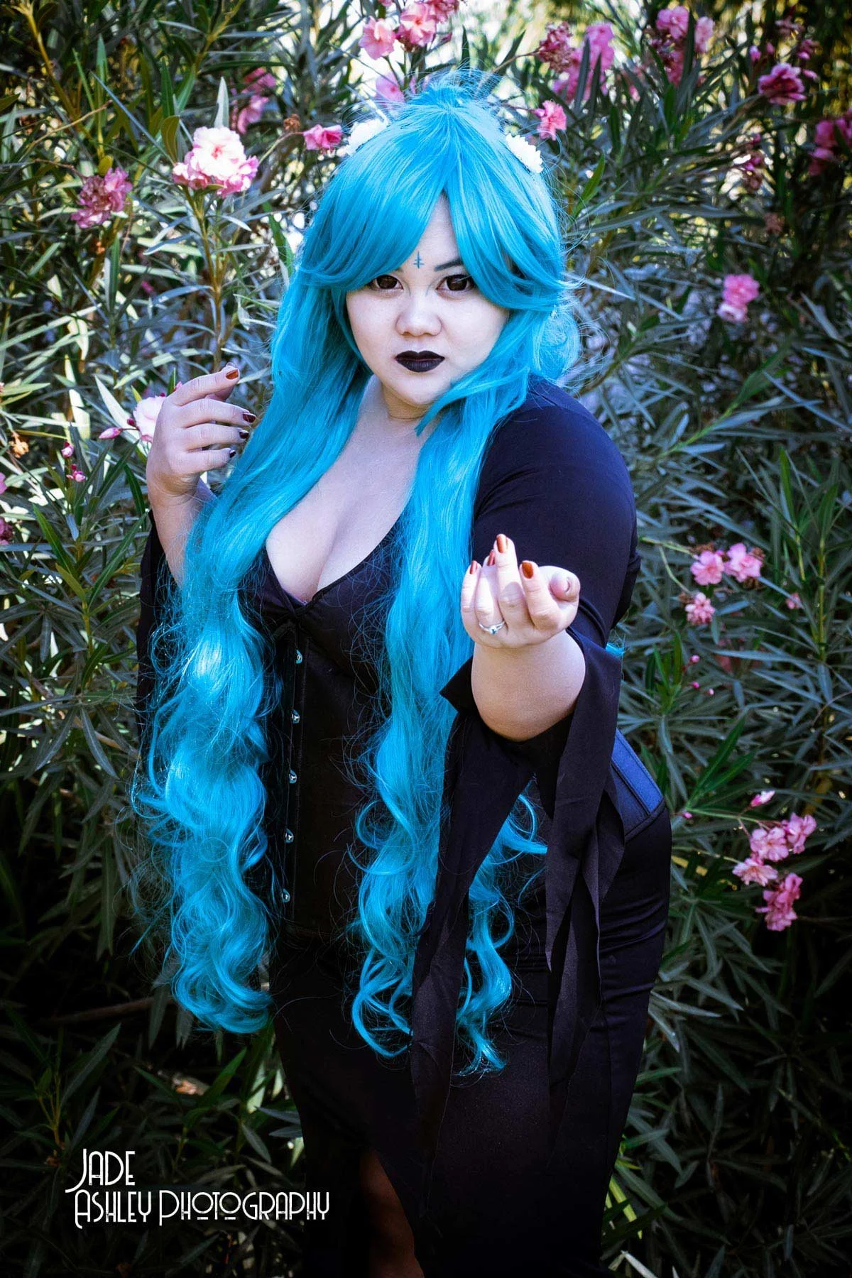 A woman with long, wavy bright blue hair and dark makeup, including black lipstick and eyeliner, stands among greenery and pink flowers. She is wearing a black top and is posing with one hand extended toward the camera.