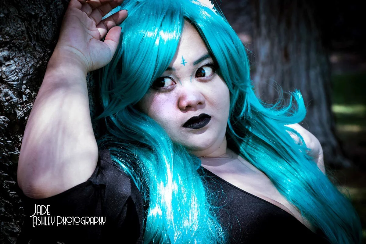 Person with bright blue hair, dark makeup, and black lipstick leaning against a tree outdoors.