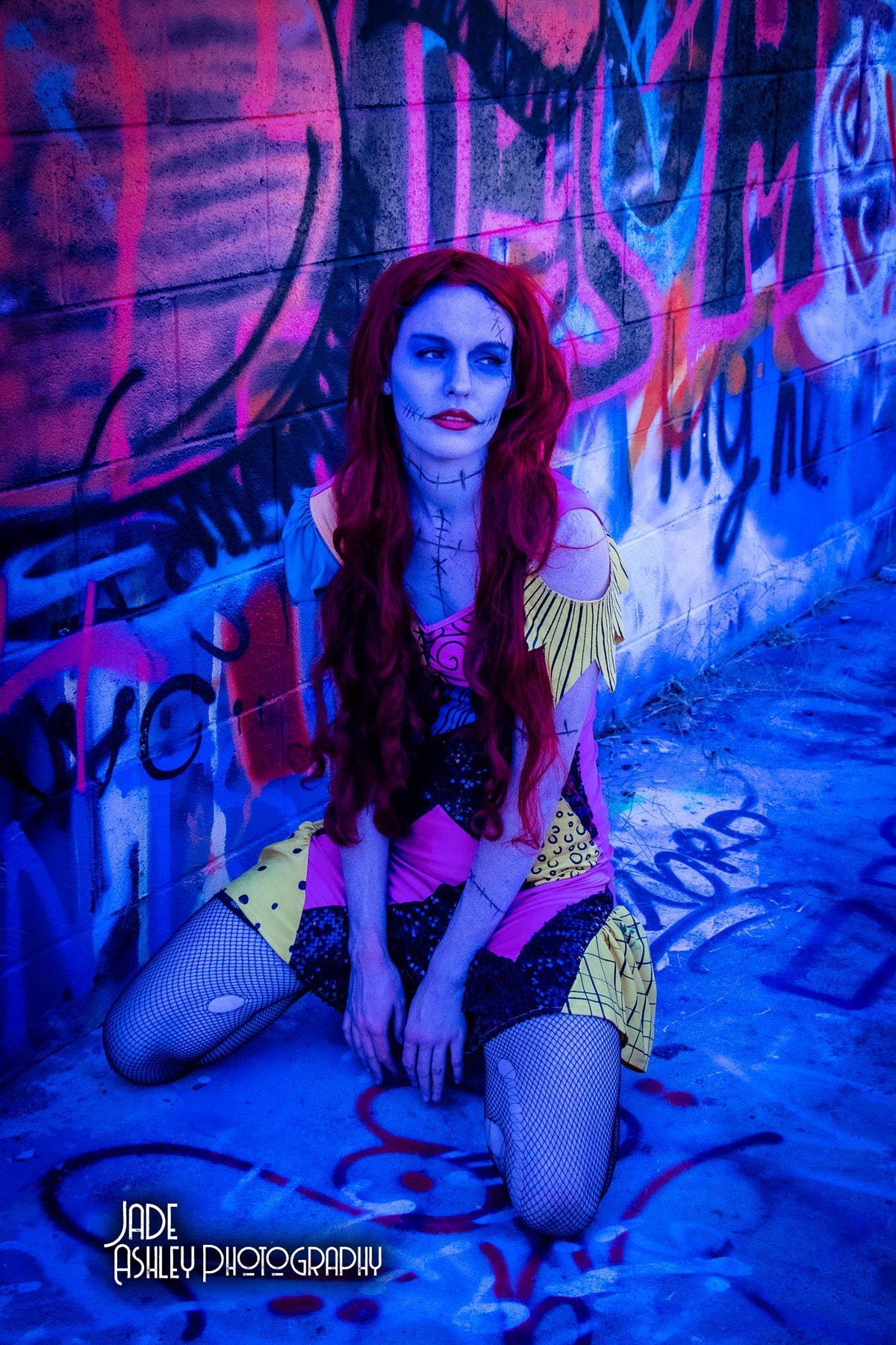 A woman with long red hair, painted blue face with stitches, sitting on the ground in front of a colorful graffiti wall, wearing a patchwork dress and fishnet stockings, illuminated with blue and pink neon lights.