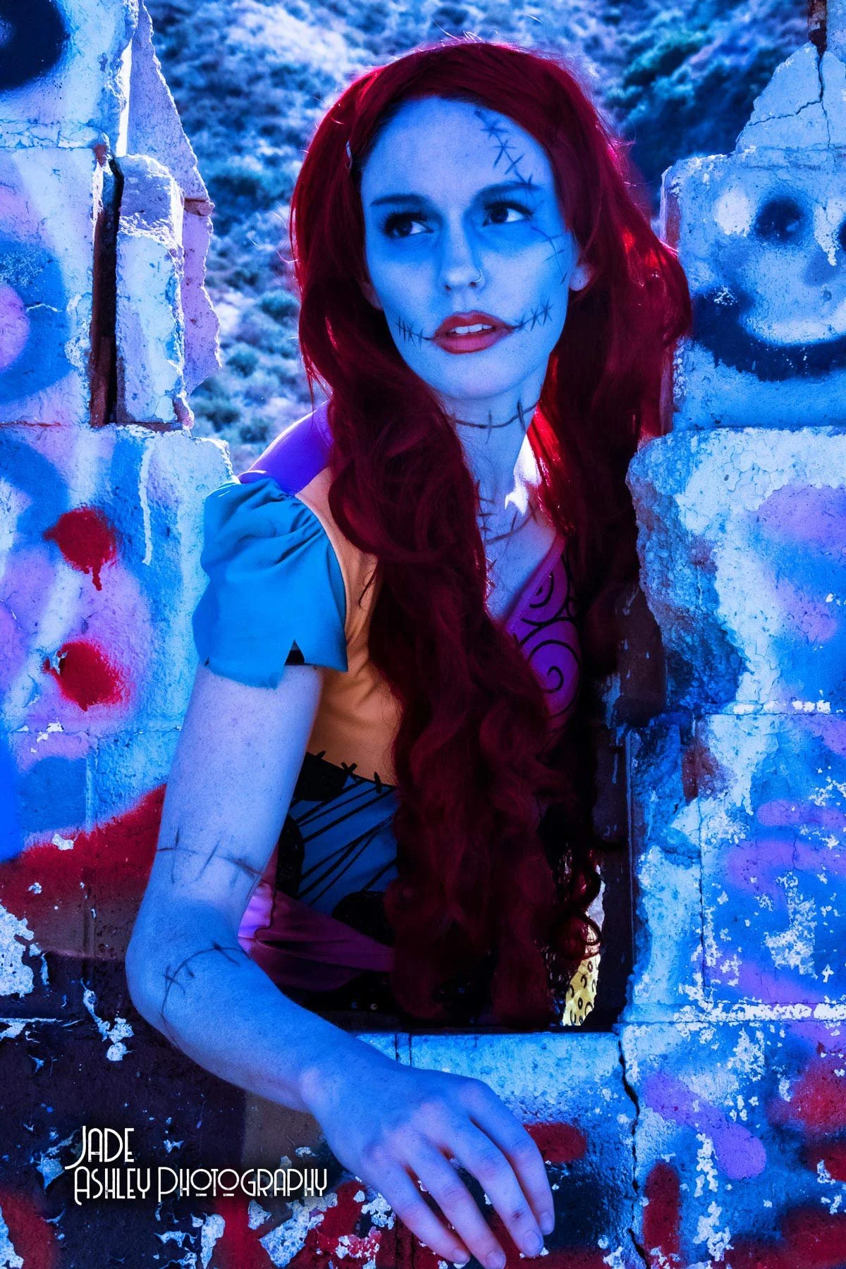 A woman dressed as a skeleton princess with red hair, wearing colorful costume, looking through a broken wall at sunset or dusk, with graffiti painted on the wall, outdoors.