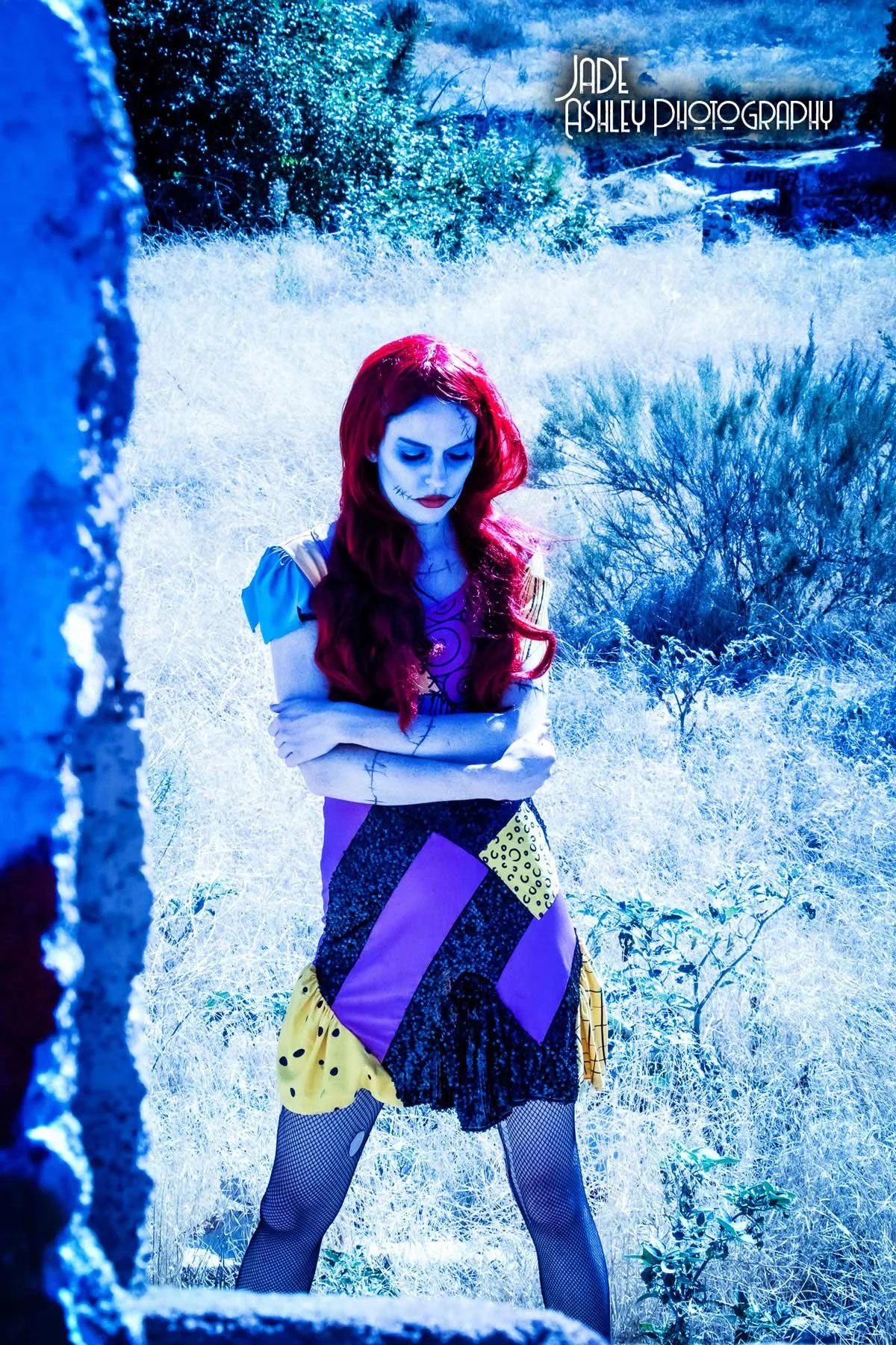 A woman dressed as a spooky character stands outdoors with her arms crossed, wearing a colorful patchwork dress, black fishnet stockings, and makeup resembling a skeleton or doll with stitches on her face and red hair.