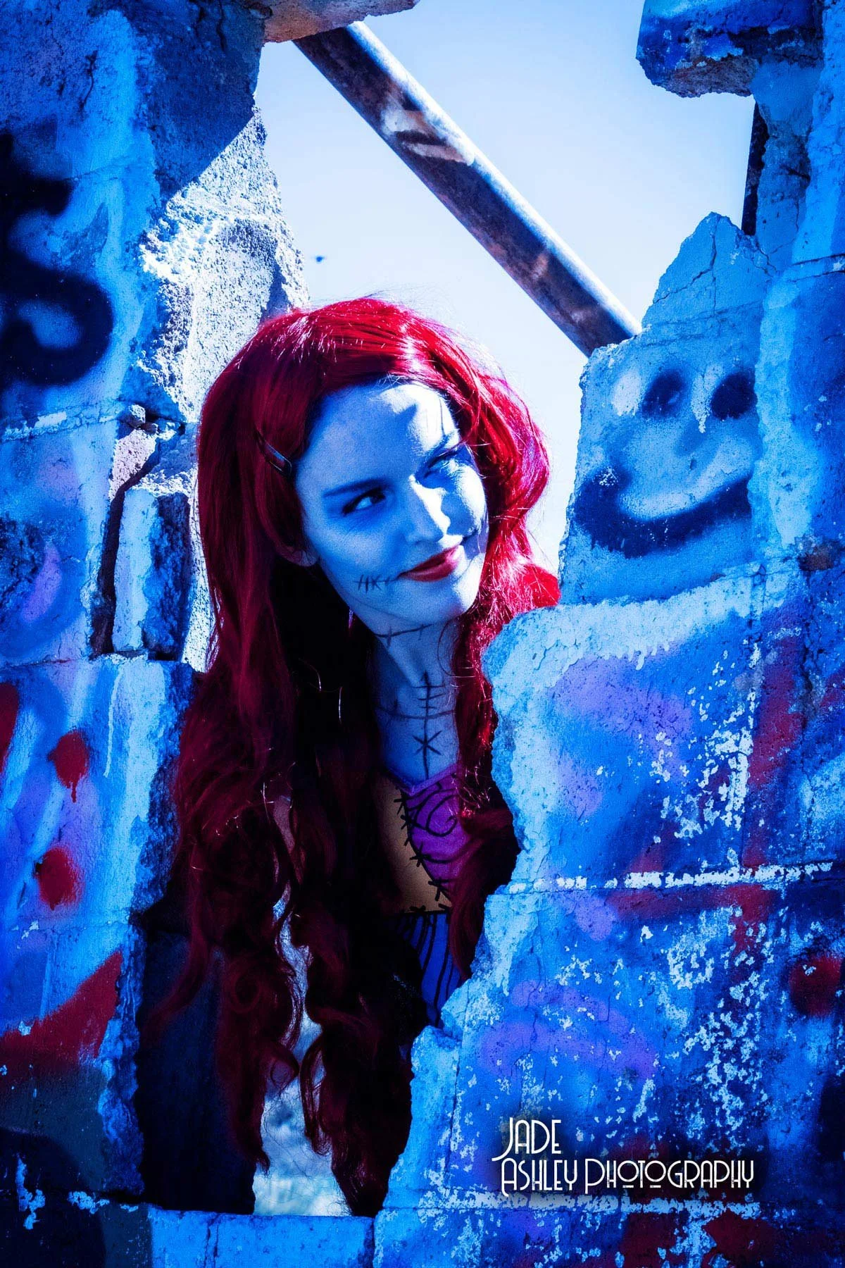 A woman with red hair dressed as a spooky character peering through a broken wall with graffiti, including a smiley face and red blood splatters, during nighttime.