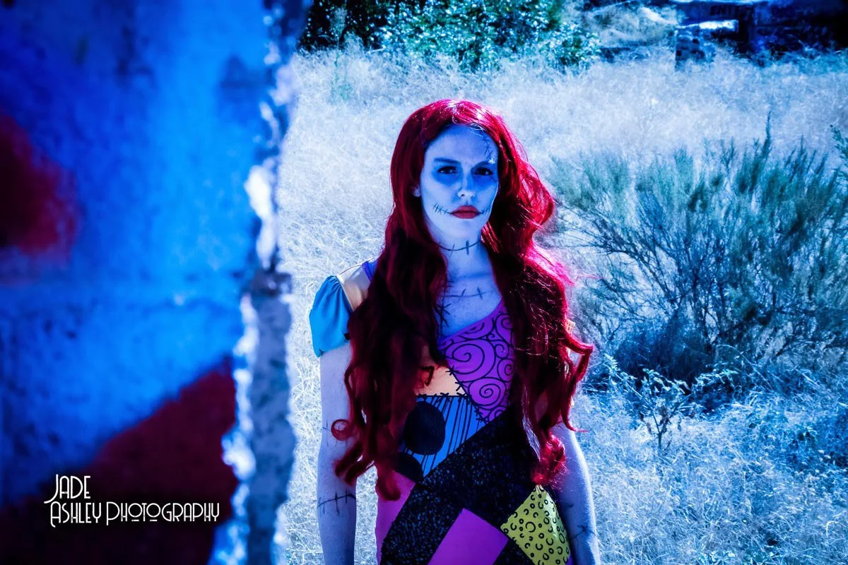 A woman dressed as a skeleton with face makeup, wearing a colorful patchwork dress, standing outdoors in a field with dry grass and bushes, partly obscured by a brick wall in the foreground.