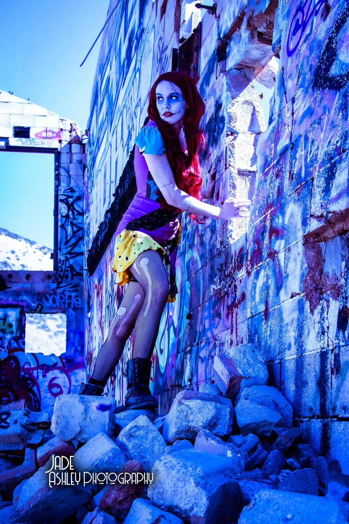 A woman dressed as a clown with a colorful costume, colorful makeup, and long red hair, stands on rocks in front of a graffiti-covered wall, holding onto the wall and looking to the side.