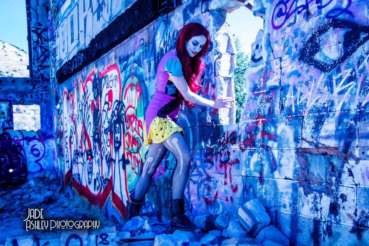 A young woman with red hair, dressed in colorful and edgy clothing, poses in front of graffiti-covered walls in an urban setting, surrounded by rubble.