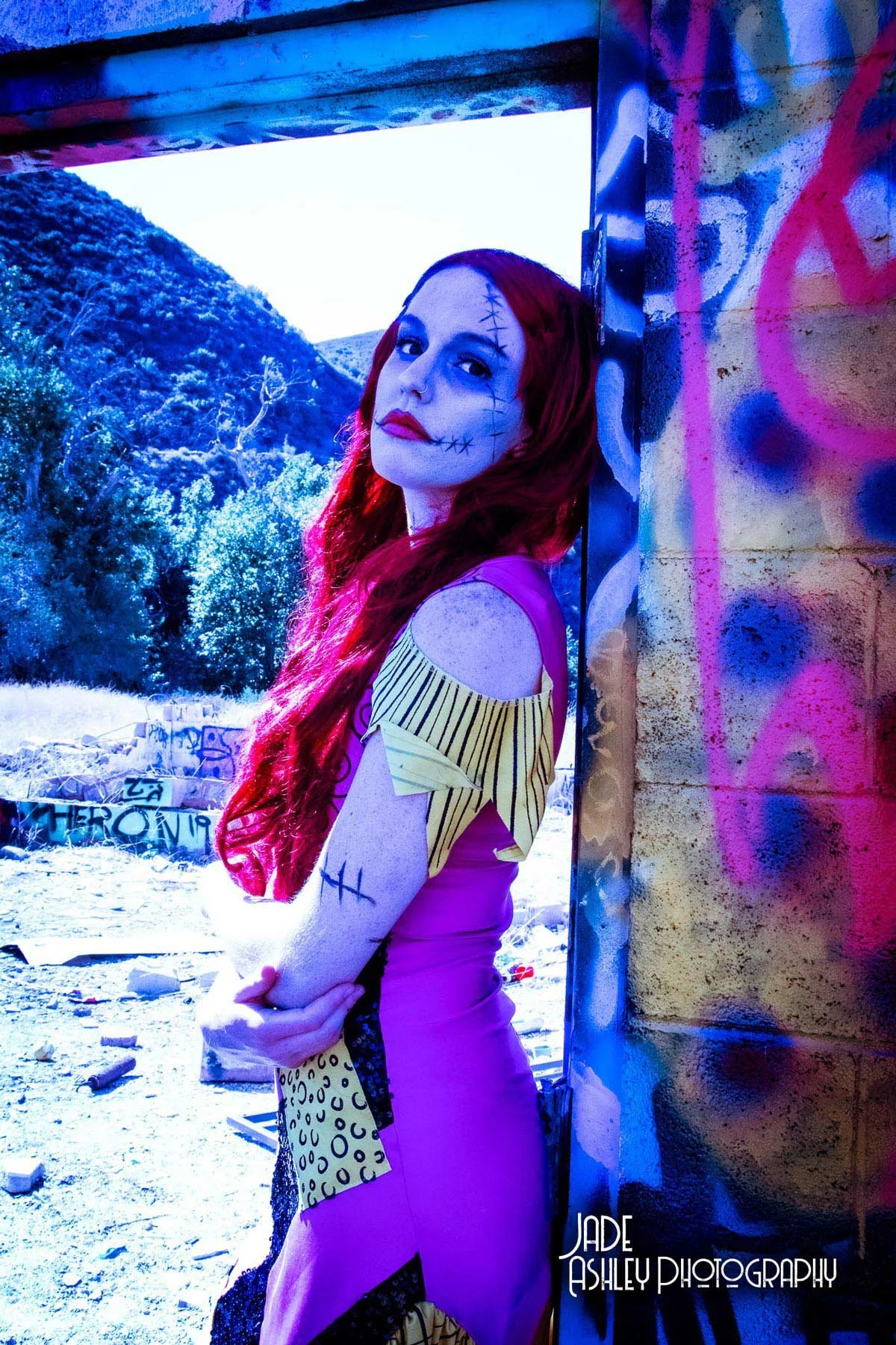 A woman with red hair and makeup resembling a sugar skull, wearing a purple dress with leopard print and yellow striped accents, leaning against a graffitied wall under an overpass with a graffiti-covered background and open landscape.