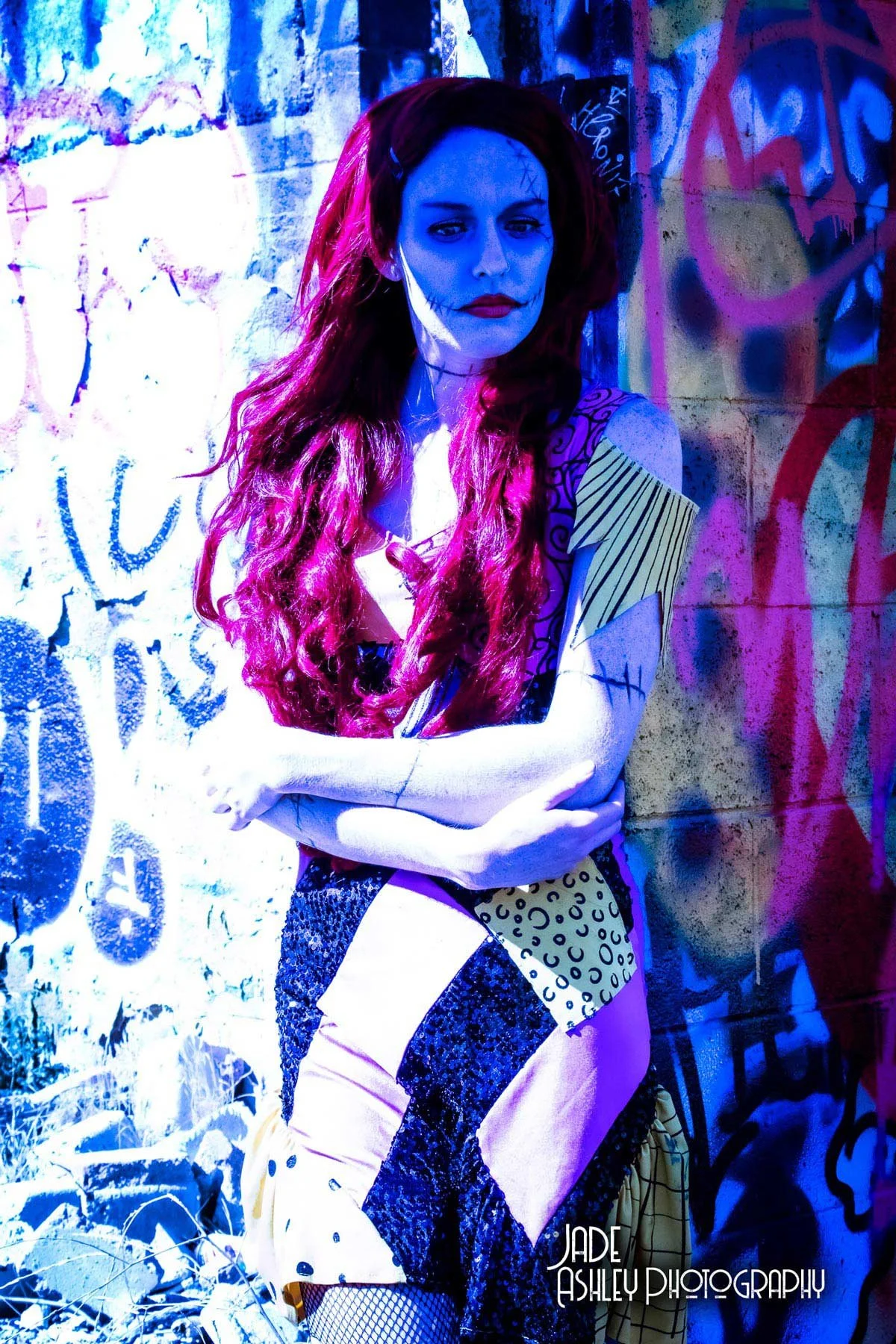 A woman with long, wavy red hair standing against a graffiti-covered wall, with her arms crossed. She has a painted face resembling a skull or day of the dead makeup, and wears a patchwork dress with various patterns. The photo is taken outdoors with