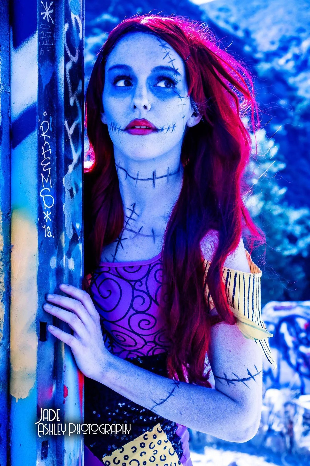 A woman with long red hair in Halloween makeup with stitches drawn on her face and body, wearing colorful clothing, standing outdoors near graffiti-covered objects with a snowy background.
