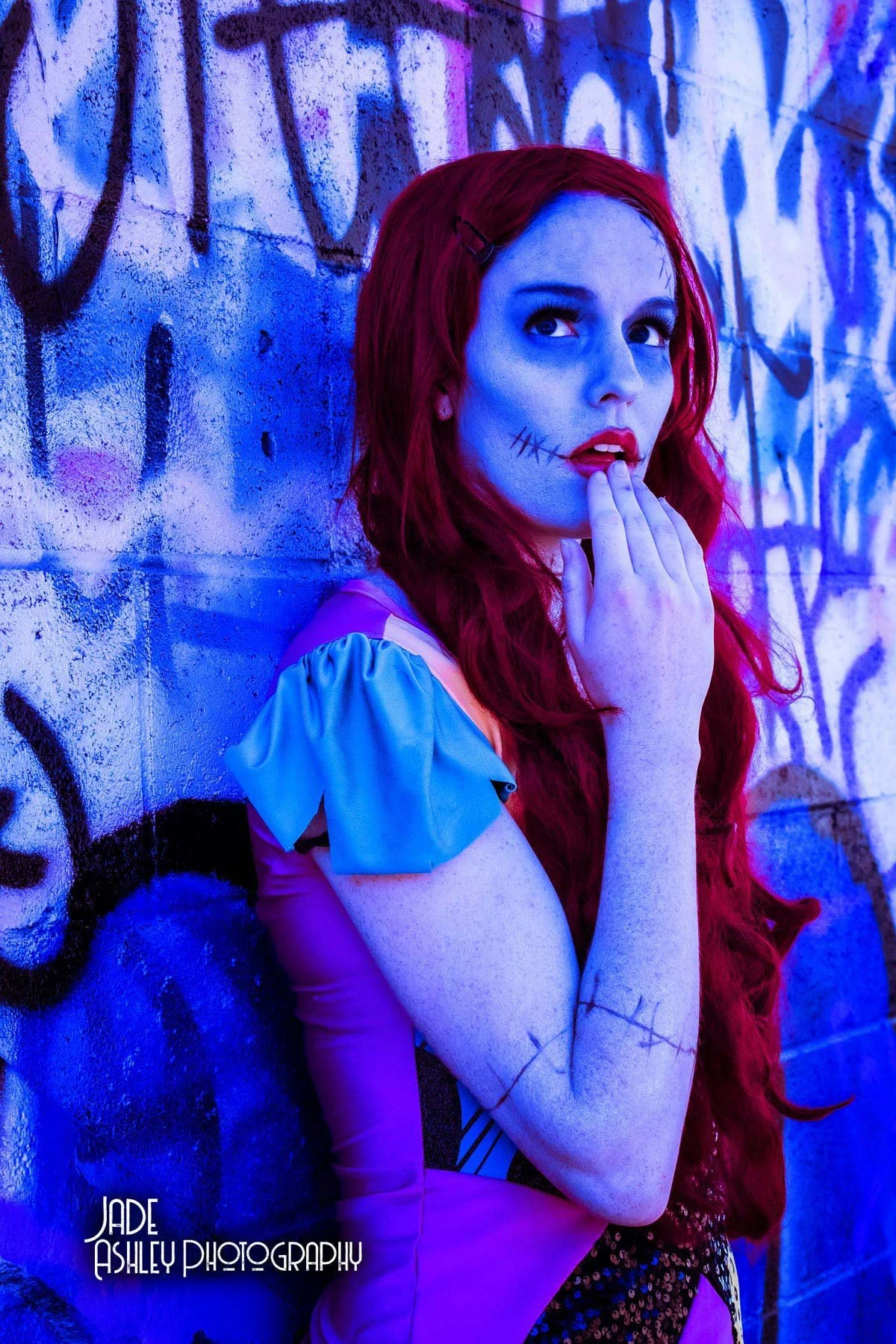 A woman with long red hair, face painted with stitched scars, stands against a graffiti-covered wall illuminated by purple and blue neon lights, wearing a costume with pastel-colored sleeves.