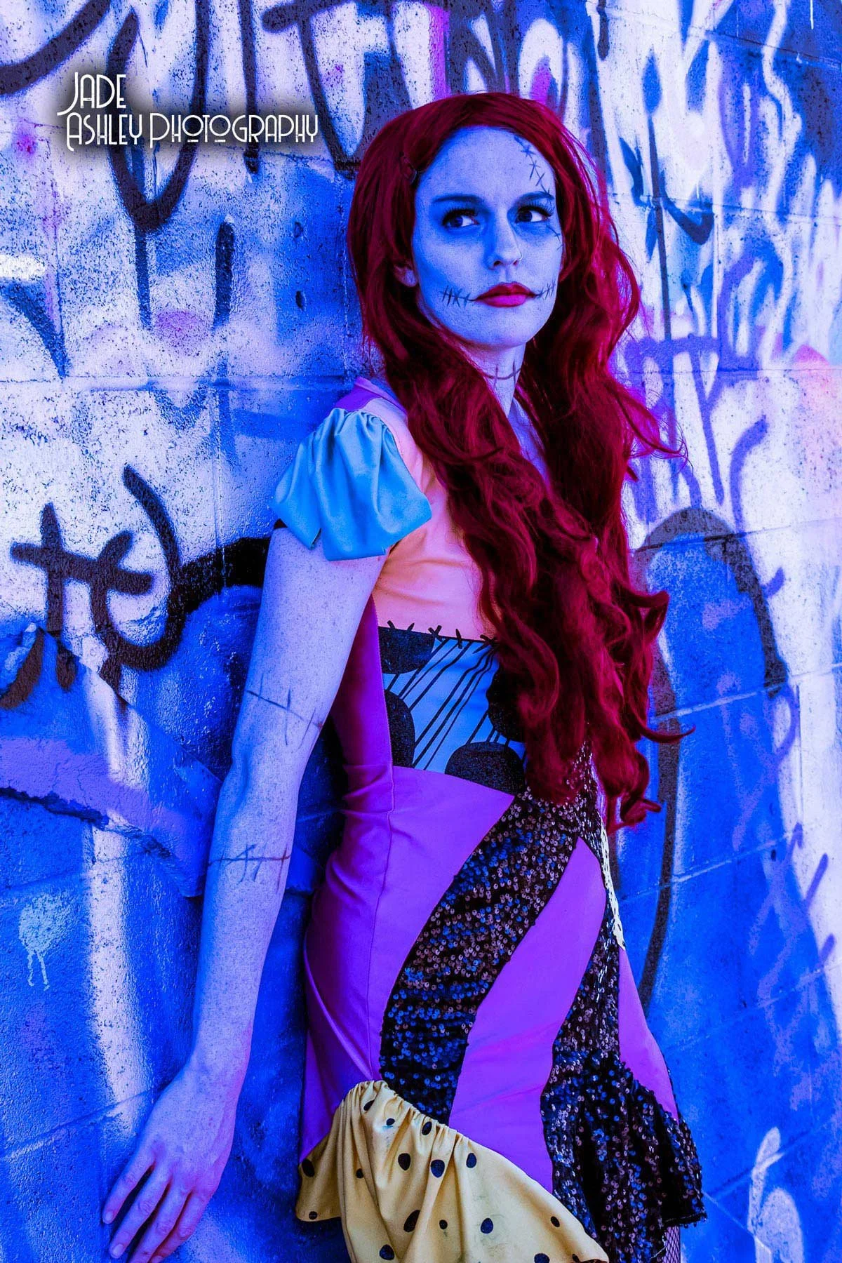 A woman dressed as a colorful clown or character with red hair and face paint showing stitches and jokey scars, standing against a graffiti-covered wall.
