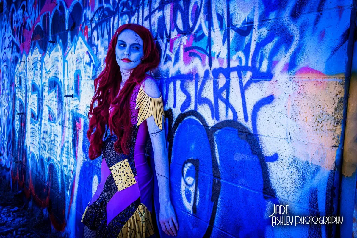 Woman dressed in colorful, patterned costume with painted face, standing against graffiti-covered wall, with blue and purple lighting.