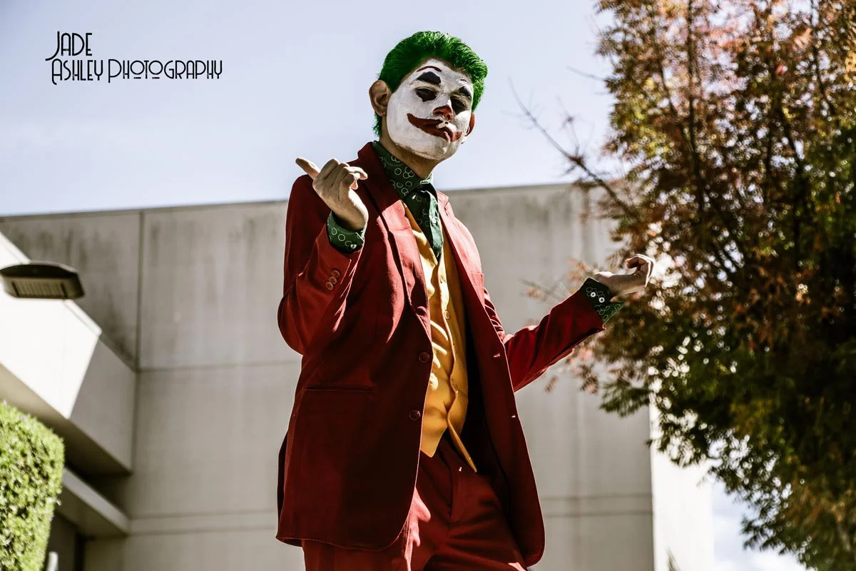 Person dressed as the Joker character from Batman, wearing a red suit, yellow vest, and green hair, standing outdoors with a large concrete building and autumn trees in the background.