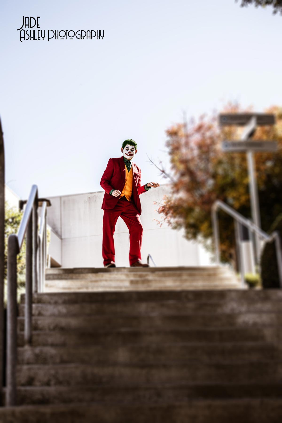 A person dressed as the Joker from Batman, wearing a red suit, orange vest, and clown makeup, standing at the top of stairs outdoors.