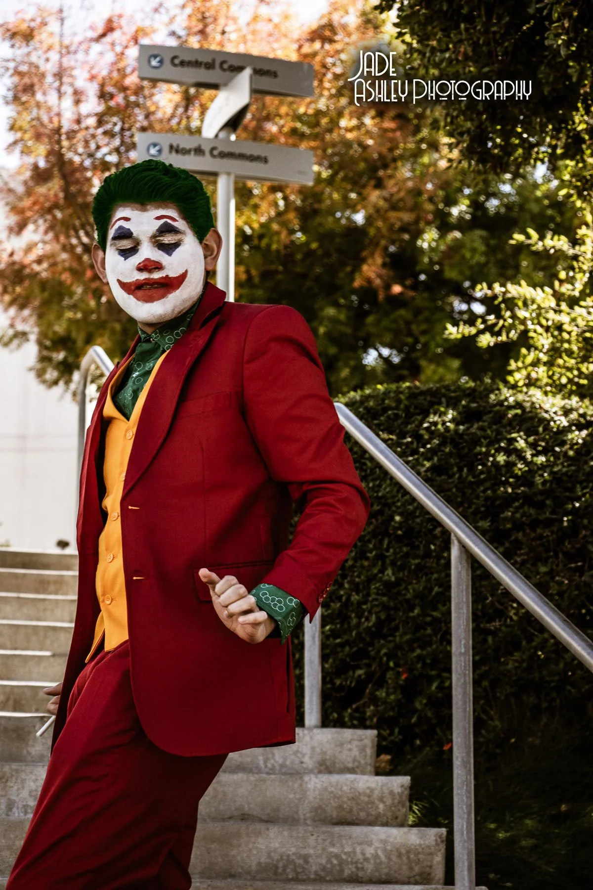A person dressed as the Joker in a red suit, yellow vest, and green shirt with patterned cuffs, standing outdoors on stairs with trees in the background and signposts pointing to Central Commons and North Commons, during daytime.