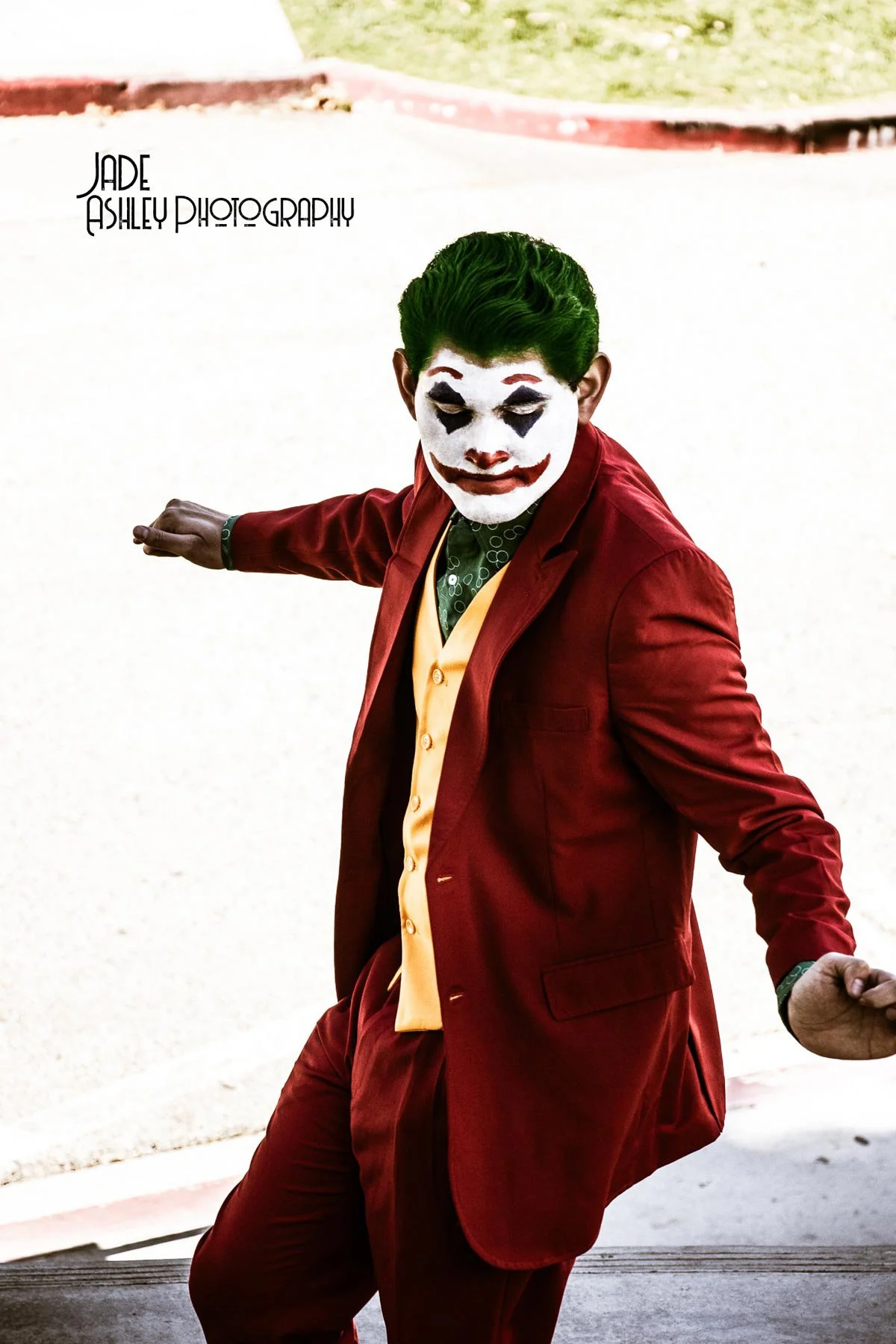 A person dressed as the Joker, wearing a red suit with a yellow vest, with green hair and clown makeup, standing outdoors on a sidewalk.