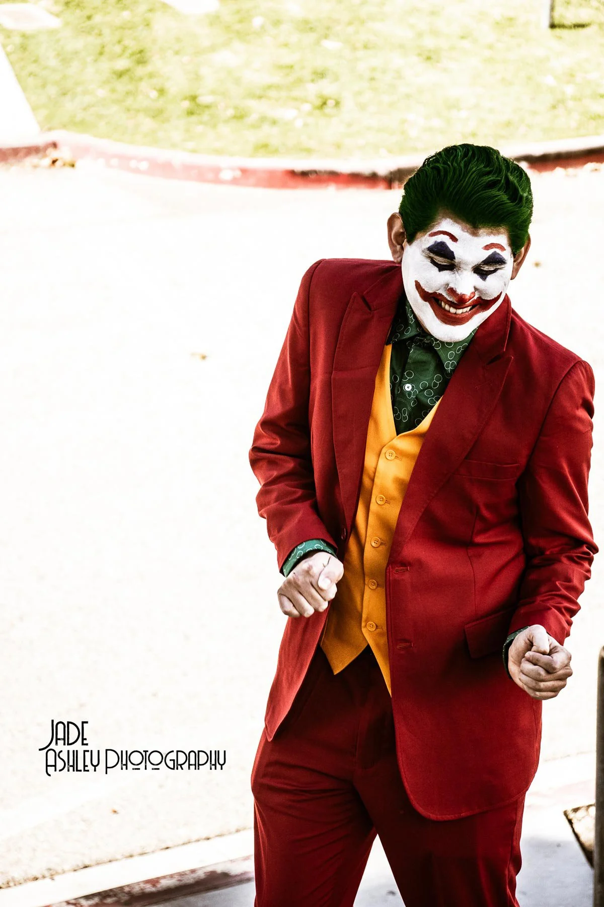 A person dressed as the Joker from Batman, wearing a red suit, yellow vest, and green shirt, with white face paint, red lips, and black eye makeup, smiling and making an animated gesture.