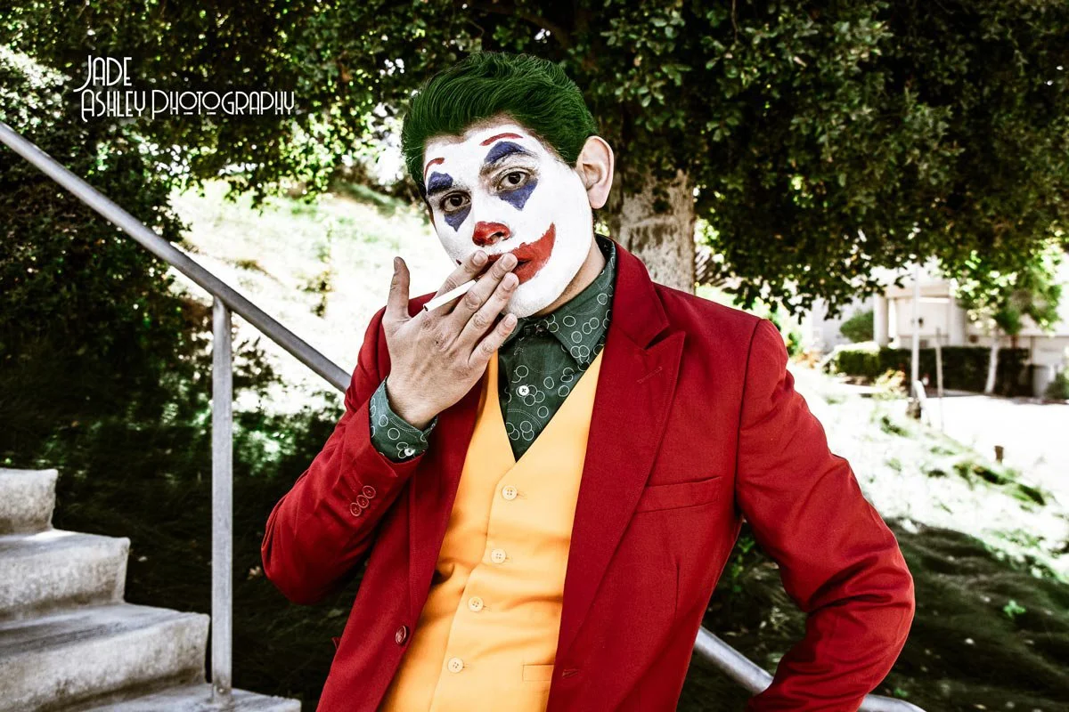 Person dressed as the Joker with clown makeup, green hair, red jacket, yellow vest, and patterned shirt, smoking a cigarette outdoors.