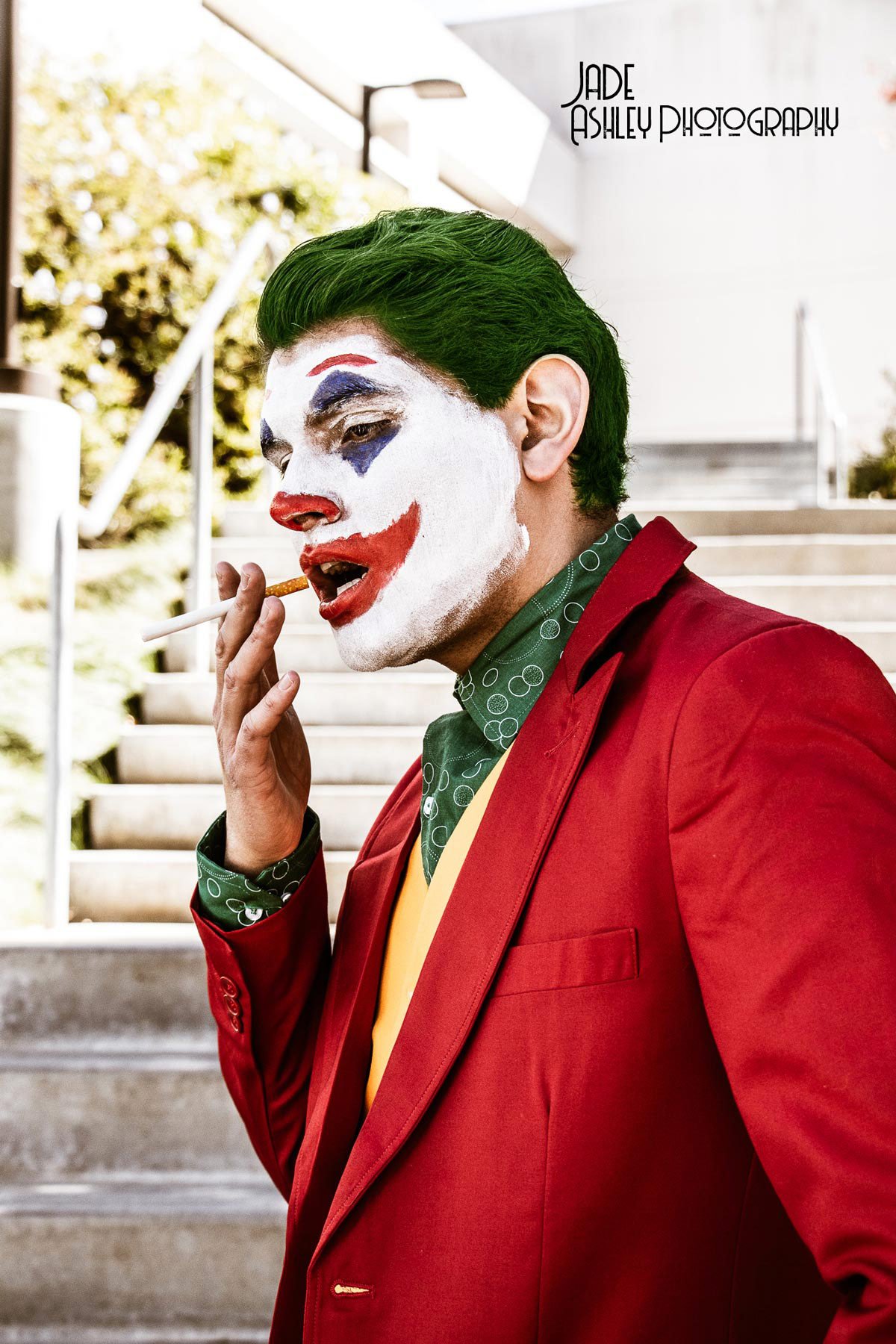 Person dressed as the Joker, with face painted white with colorful clown makeup, wearing a red jacket, green shirt, and yellow tie, smoking a cigarette outdoors near a staircase.