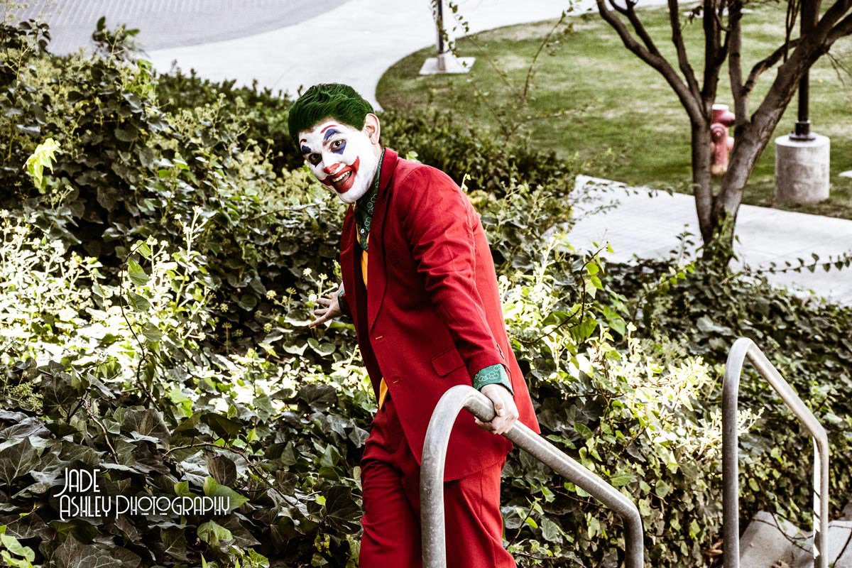 A person dressed as the Joker with clown makeup and red suit climbing up outdoor stairs surrounded by greenery.