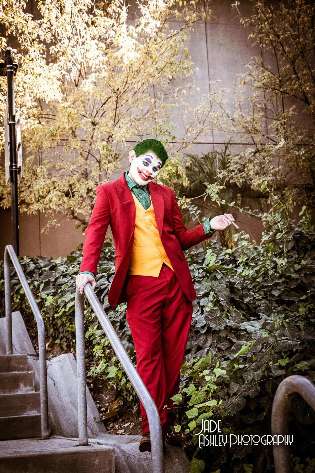 A person dressed as the Joker from Batman, wearing a red suit with a yellow vest and green shirt, standing on outdoor stairs with a metal railing, surrounded by trees and foliage.