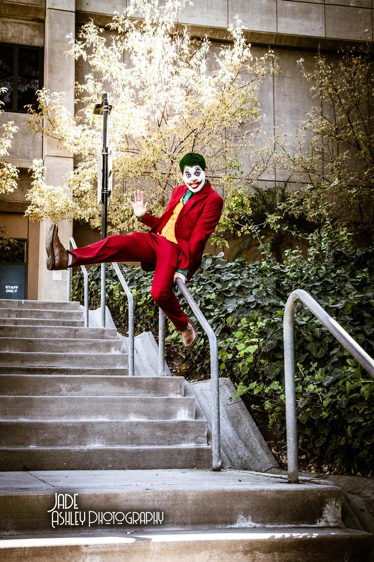 A person dressed as the Joker from Batman, wearing a red suit, yellow vest, green shirt, and clown makeup, sitting on a handrail at the top of concrete stairs outdoors with greenery and trees in the background.