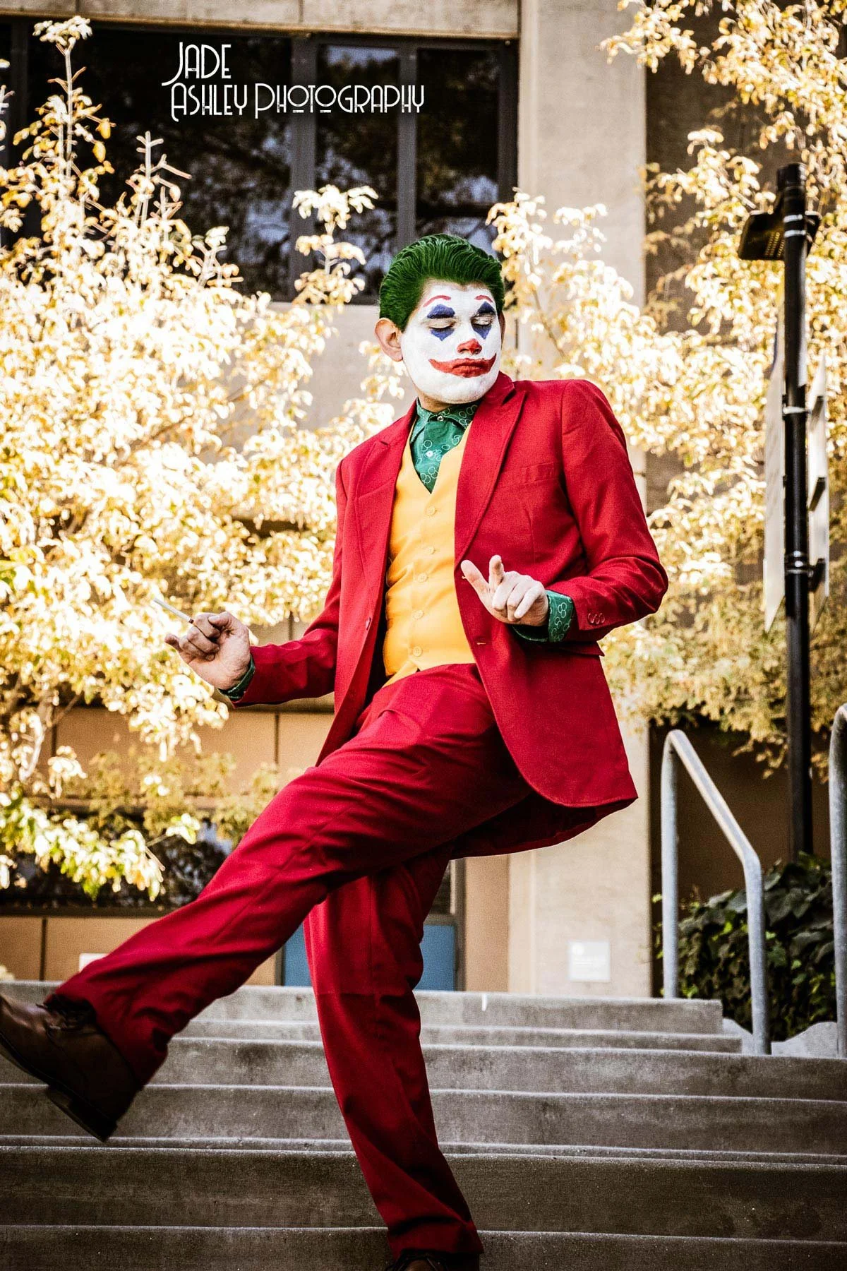A person dressed as the Joker in a red suit, yellow vest, and green shirt, with clown makeup, standing on outdoor stairs with autumn trees in the background