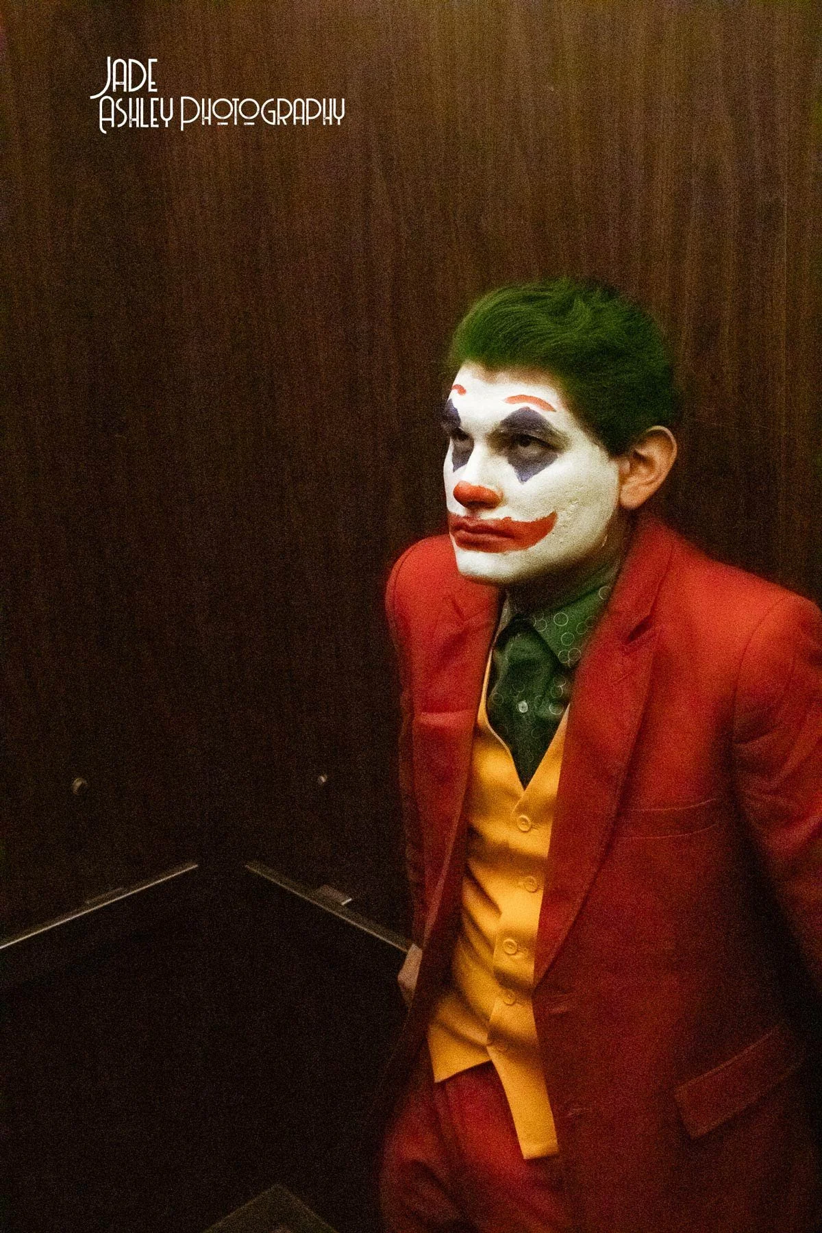 Person dressed as the Joker in an elevator with dark wood walls, wearing a red suit, yellow vest, green shirt, and clown makeup with green hair.