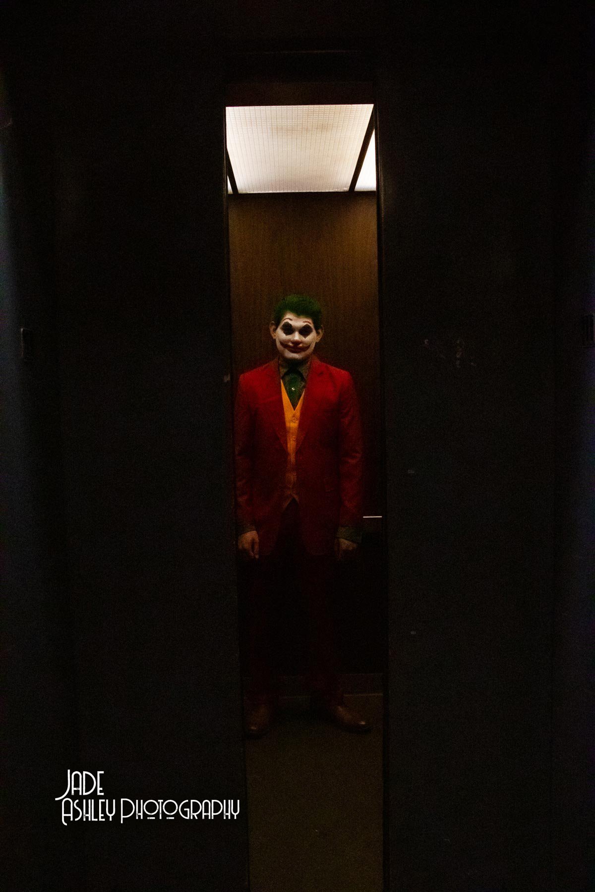 A person dressed as the Joker with face paint, green hair, red suit, yellow vest, and green shirt, standing in a narrow elevator with dark walls and a lit ceiling.