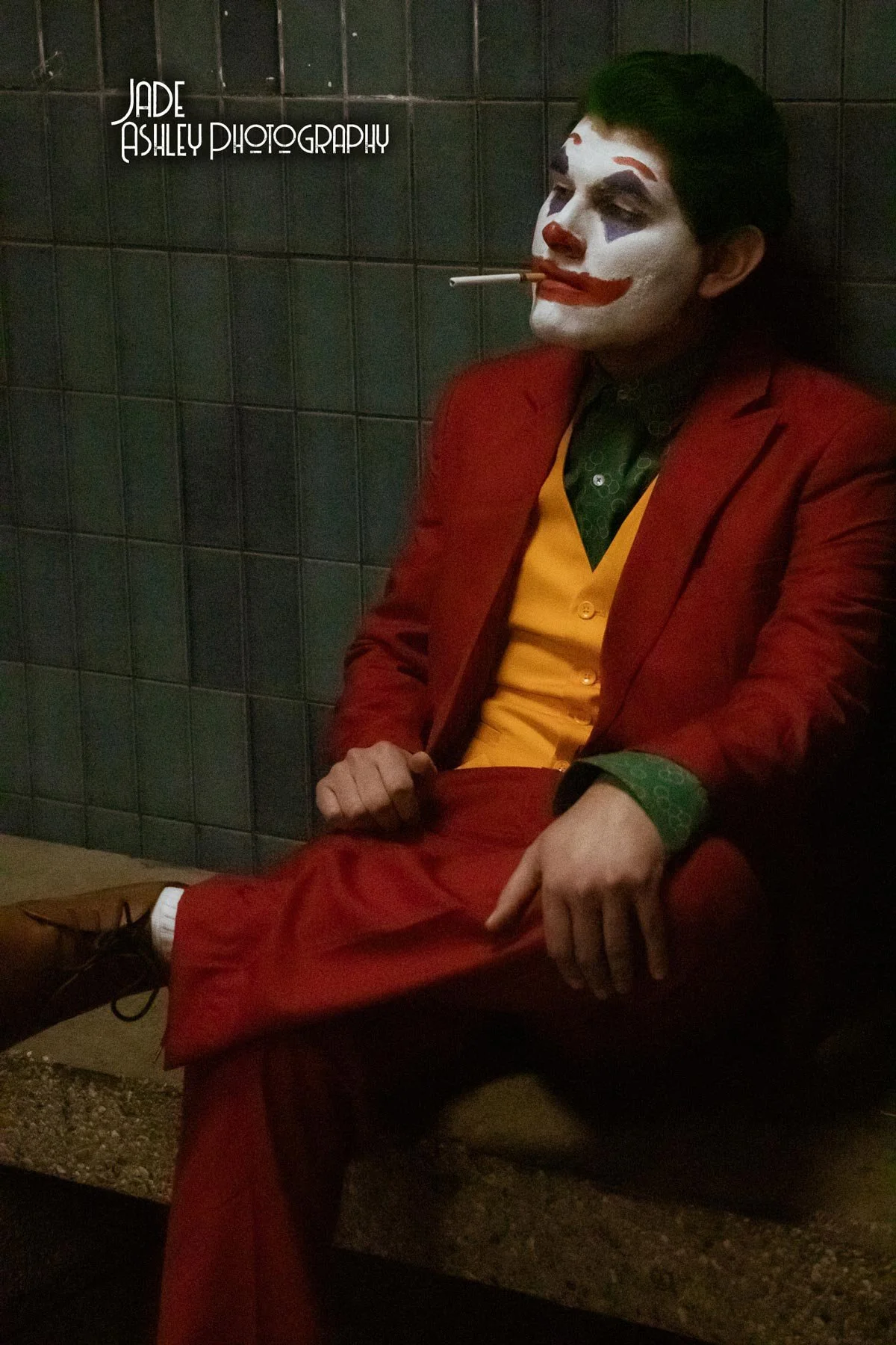 Man dressed as Joker sitting against a tiled wall with face painted in clown makeup, cigarette in mouth, wearing a red suit, yellow vest, and green shirt.