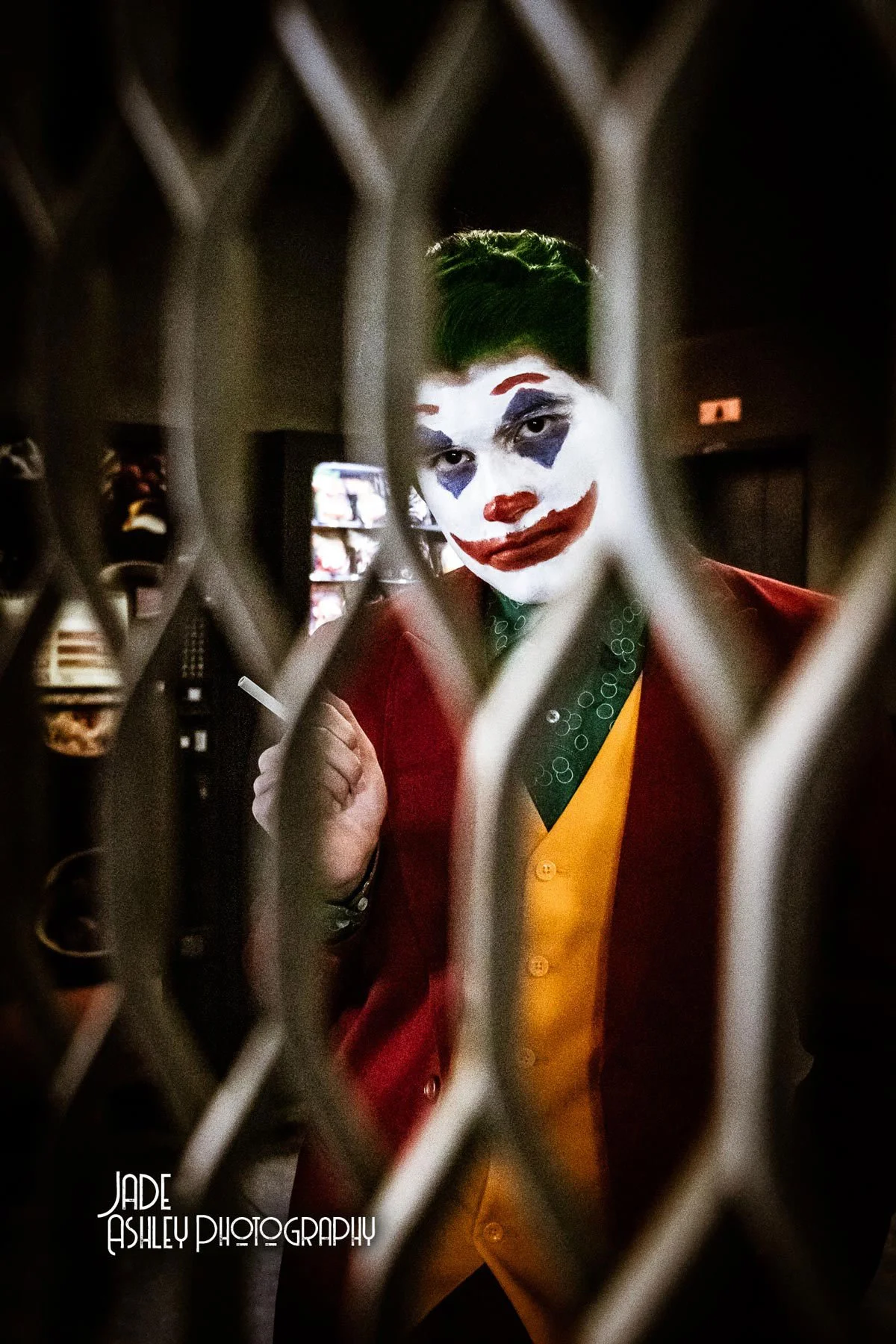 Person dressed as the Joker with clown makeup and green hair, seen through a metal fence.