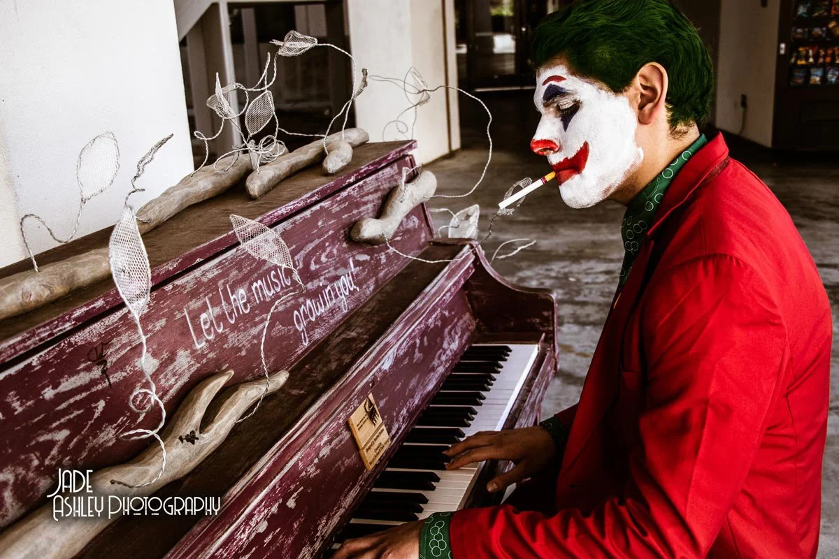 A person dressed as a clown with green hair, white face paint, and a red suit playing a worn, red piano decorated with white wire and fabric leaves, with the phrase "Let the music grow on you" written on it.
