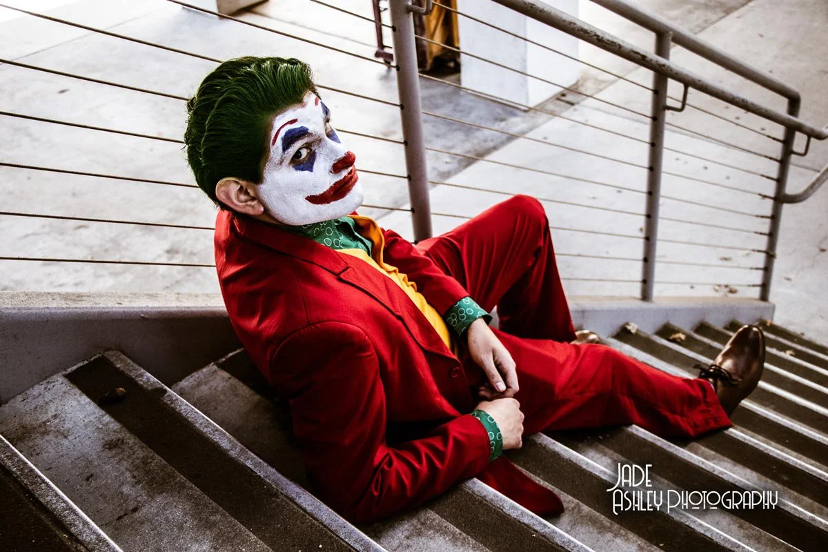 A person dressed as the Joker in a red suit with face paint, sitting on a staircase and looking at the camera.