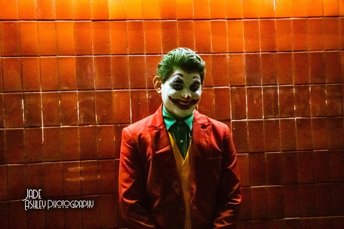 A person dressed as the Joker from Batman in a red jacket, yellow vest, green shirt, with clown makeup, smiling and standing against a tiled wall.