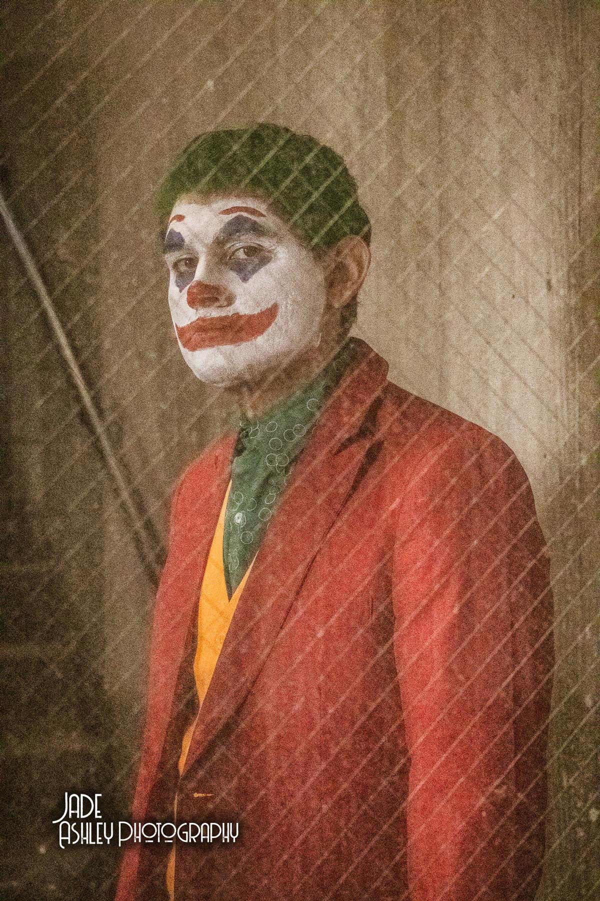 A person dressed as the Joker, with clown makeup and a red suit, looking through a mesh or glass surface.
