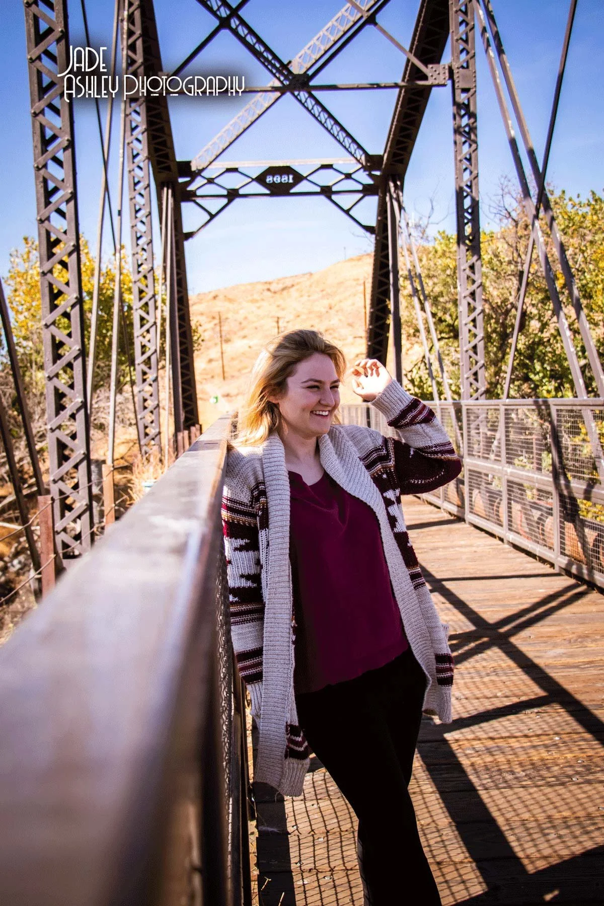 A young woman with blonde hair smiling on a metal bridge, wearing a sweater and a maroon shirt, with trees and a blue sky in the background.