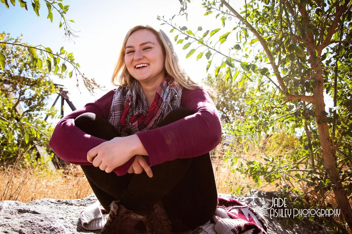 Smiling young woman sitting outdoors on a rock with trees and sunlight in the background.