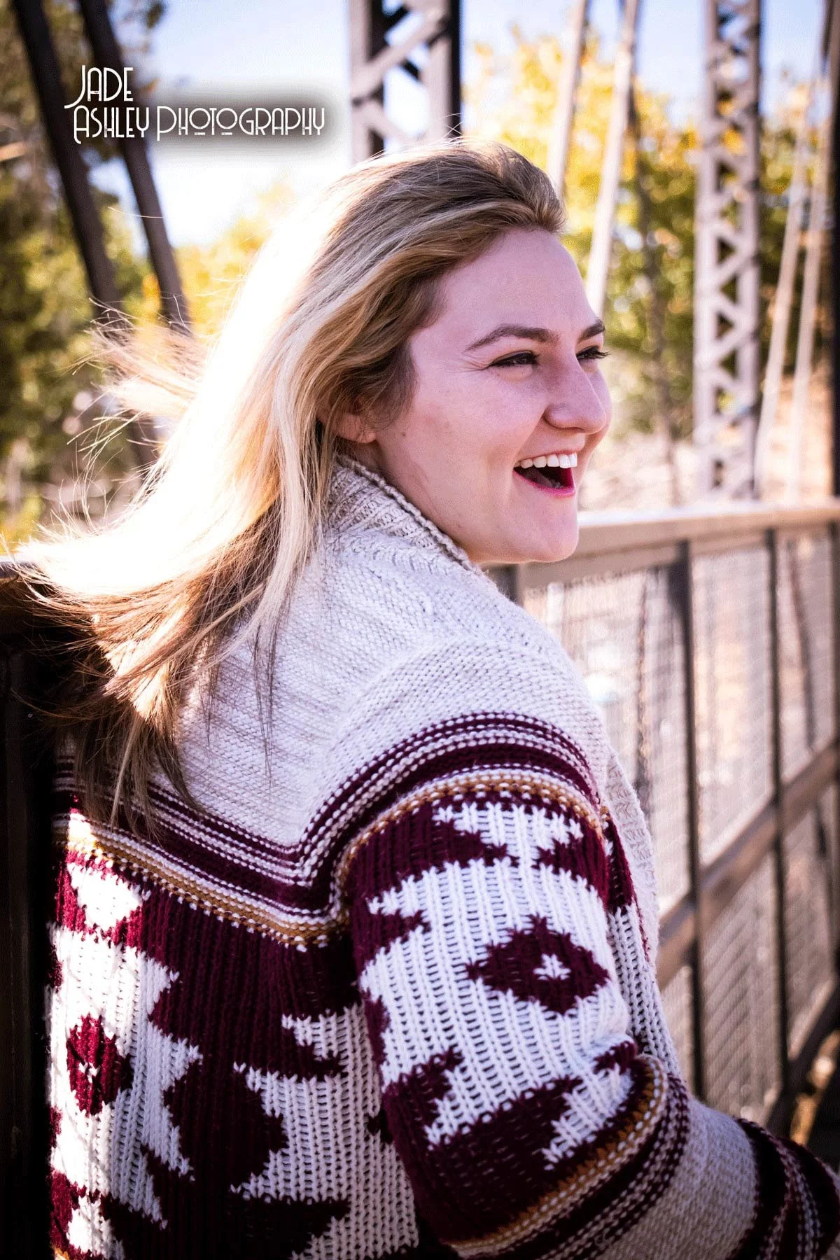A young woman with blonde hair smiling and looking to the side outdoors on a bridge during fall.