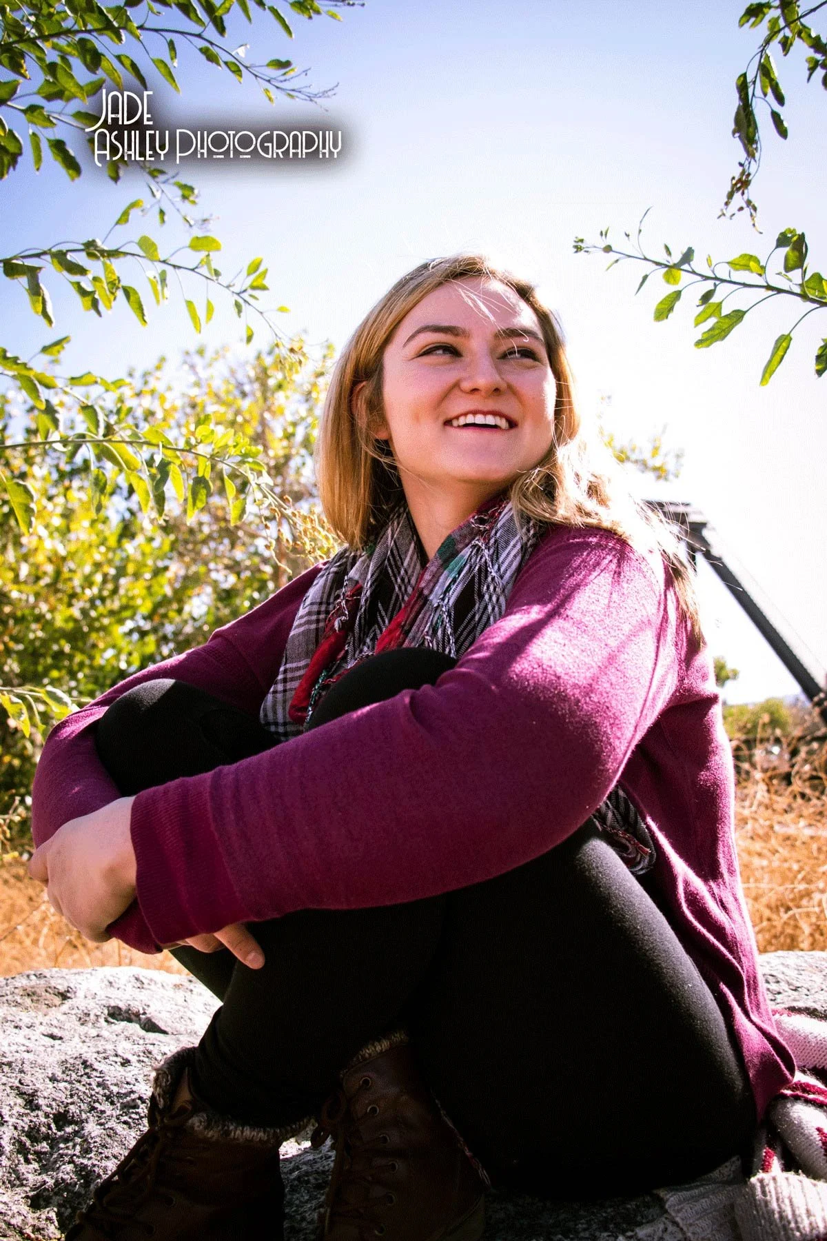 A young woman with light brown hair, wearing a burgundy sweater, plaid scarf, and brown boots, sitting on a rock outdoors surrounded by trees with sunlight and blue sky.