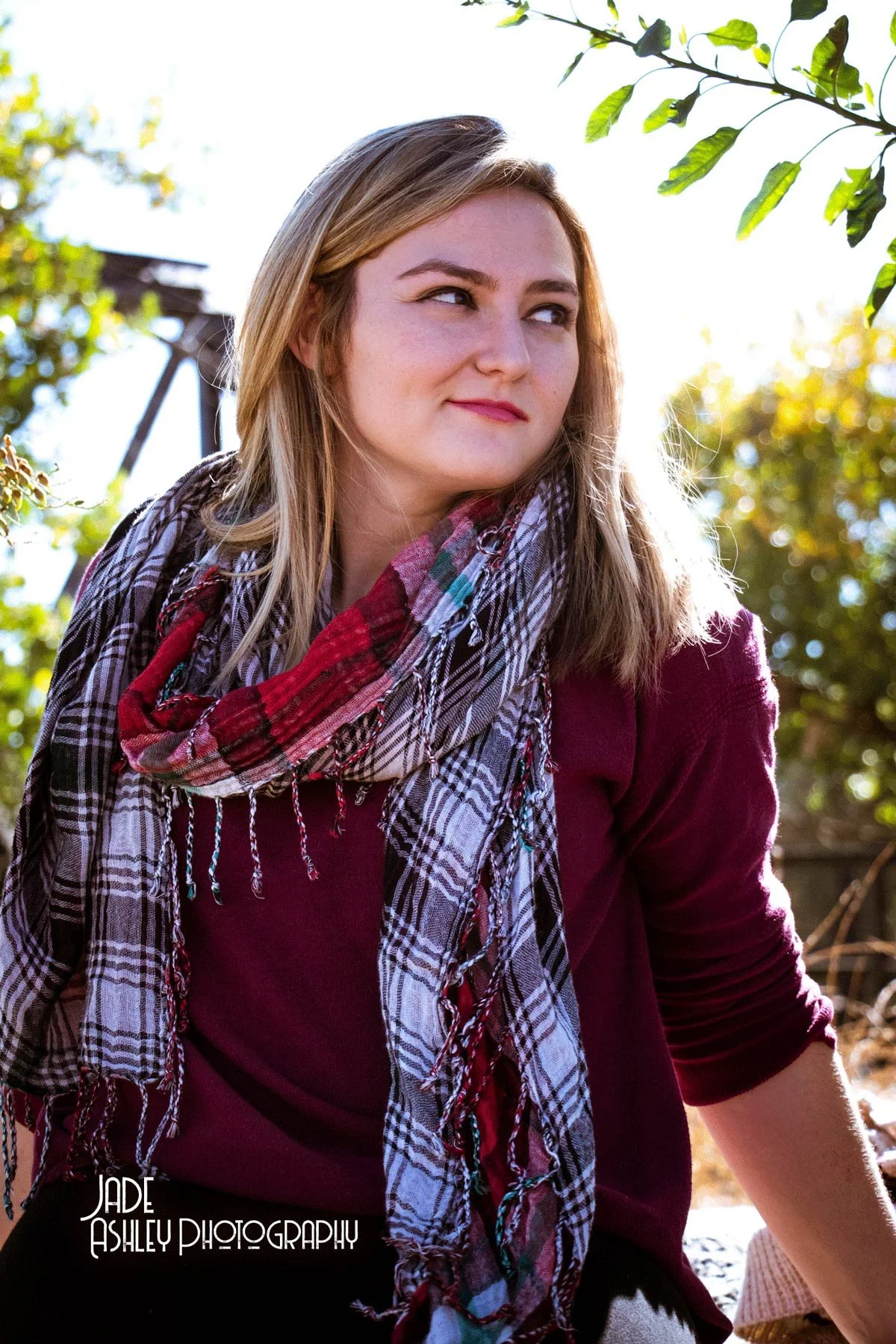 A woman with blonde hair wearing a maroon top and a red, black, and white plaid scarf, sitting outdoors surrounded by greenery and sunlight.