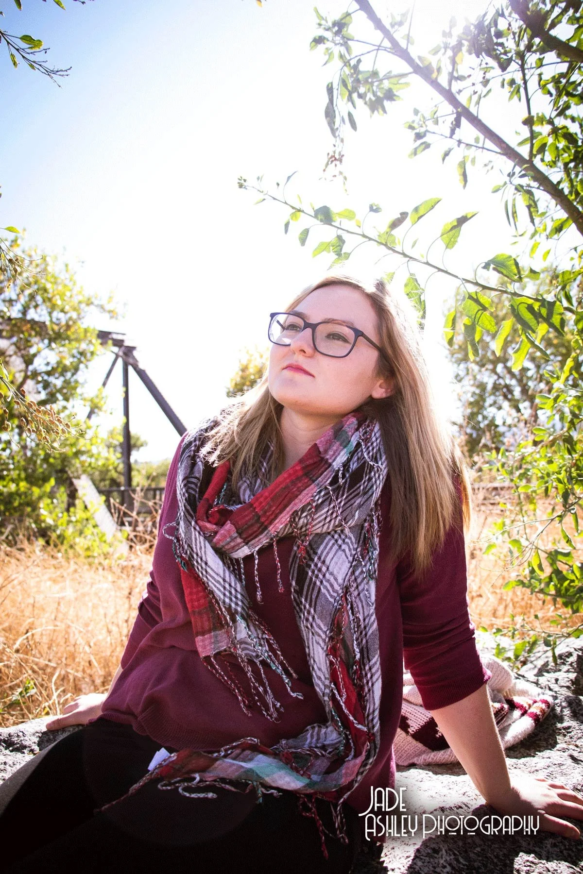 Young woman with glasses, wearing a red sweater and a plaid scarf, sitting outdoors in a sunny, natural setting with trees and dry grass.