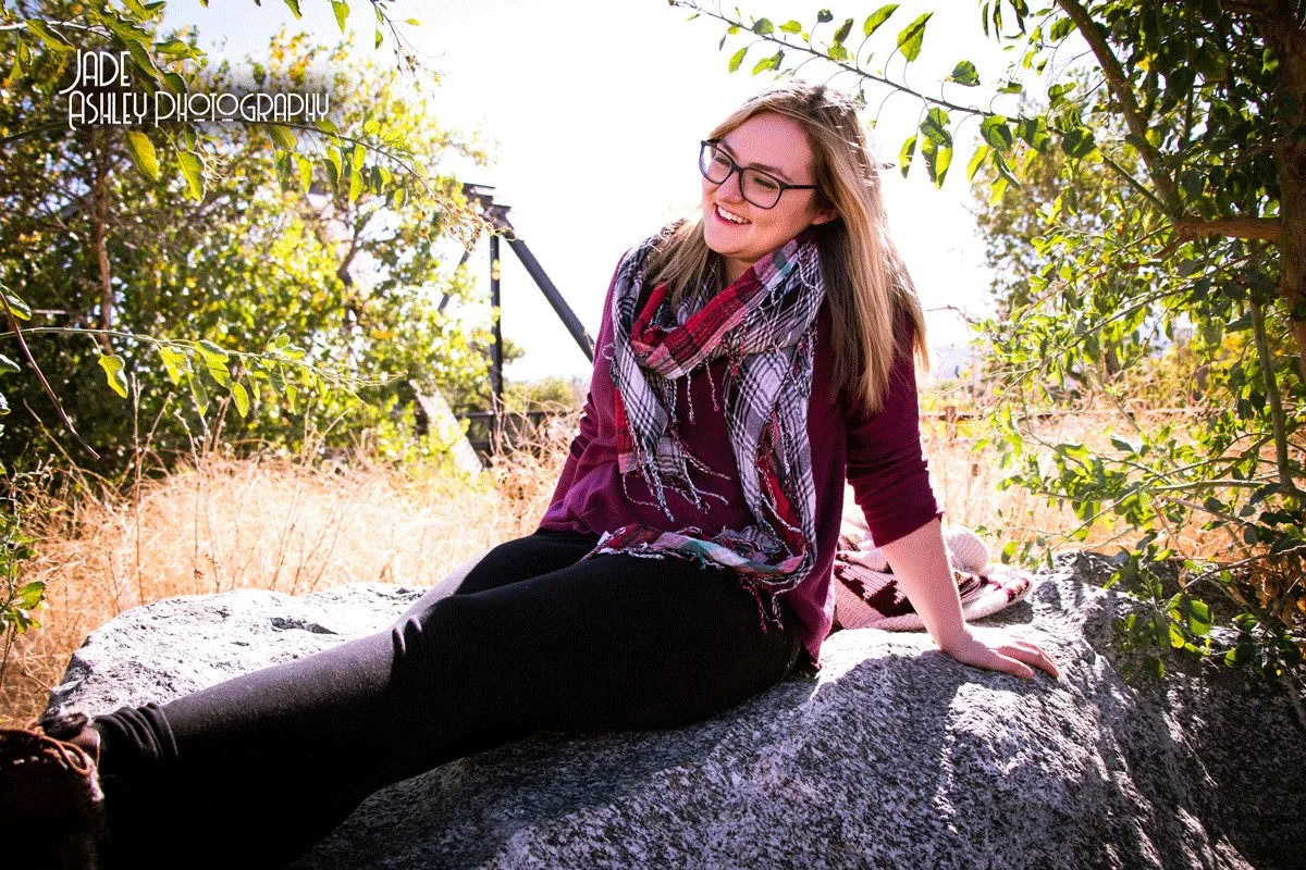 A young woman sitting on a large gray rock outdoors, smiling, with trees and sunlight in the background, wearing glasses, a purple top, black pants, and a patterned scarf.