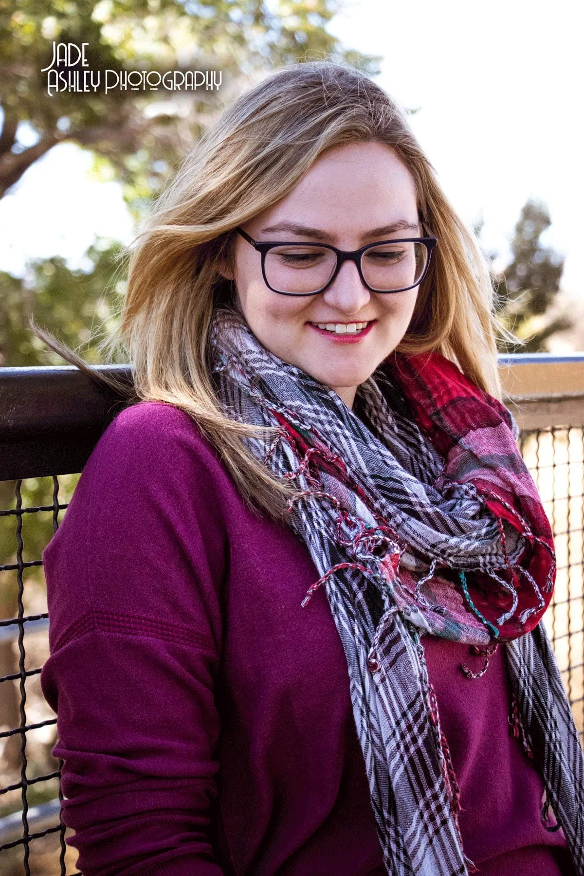 Young woman with glasses smiling outdoors, wearing a purple sweater and a plaid scarf, standing in front of a black metal fence with trees in the background.