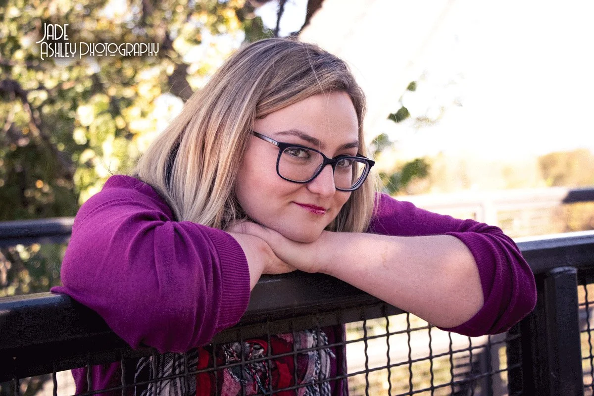 Young woman with glasses leaning on a metal fence outdoors, smiling softly, wearing a purple top, with blurred green trees in the background.
