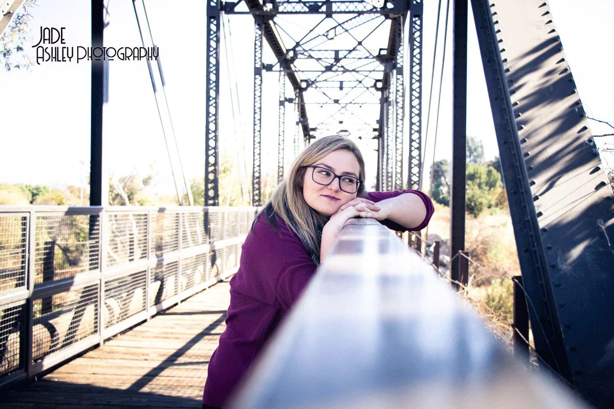 A young woman with glasses and long blonde hair leaning on a bridge railing, looking thoughtfully into the distance, with a metal bridge structure overhead and a clear sky in the background.