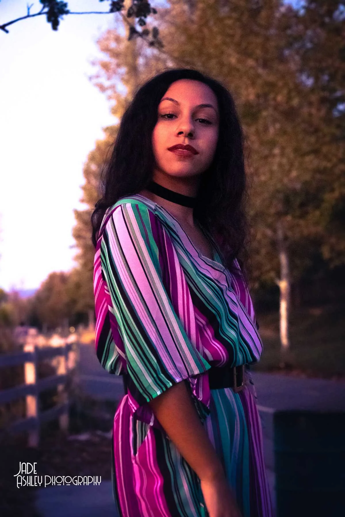 A woman with long, curly dark hair standing outdoors during sunset, wearing a colorful striped dress and a black choker, looking confidently at the camera.