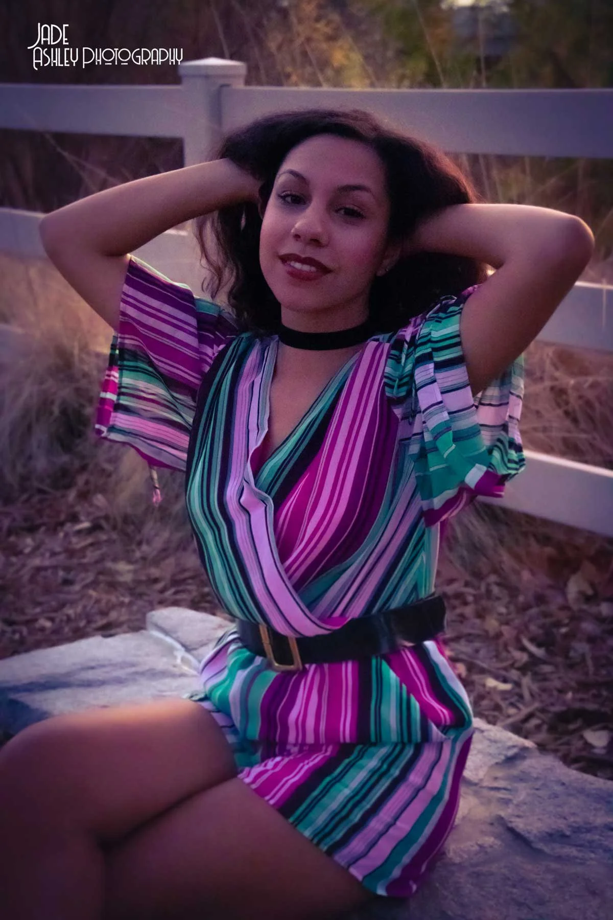 A young woman with dark hair and light skin wearing a colorful striped dress and black choker, sitting outdoors on a stone surface with her arms behind her head, smiling at the camera. There is a wooden fence and dry grass in the background, and the 