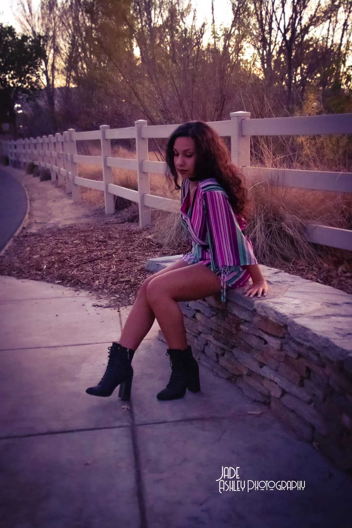 A woman with long, curly hair sitting on a stone ledge next to a sidewalk, wearing a colorful striped shirt and black high-heeled boots, during sunset in a park with trees and a white fence.