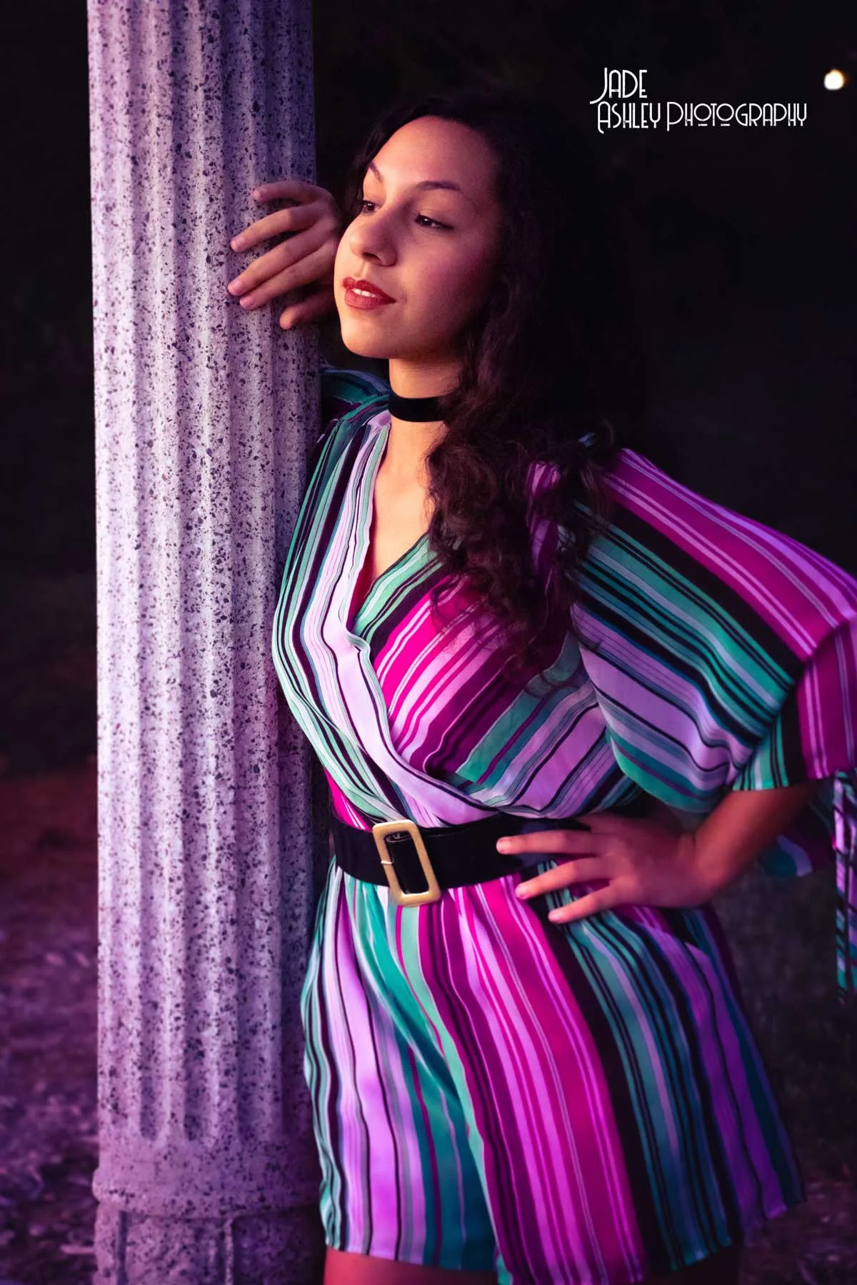 A woman with dark curly hair poses next to a textured white column at sunset, wearing a colorful striped dress with a black belt and a choker, with a confident expression.